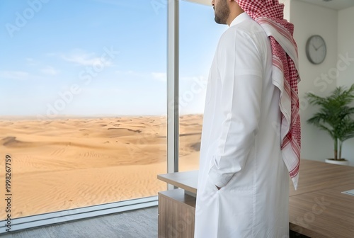Man wearing a thobe and ghutra, standing by a large glass window overlooking a vast sandy desert landscape at sunset.