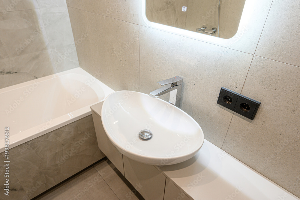 Fototapeta premium modern bathroom with a white oval sink on a sleek countertop, built-in bathtub, illuminated mirror, chrome faucet, neutral marble-textured tiles, dual power outlets.