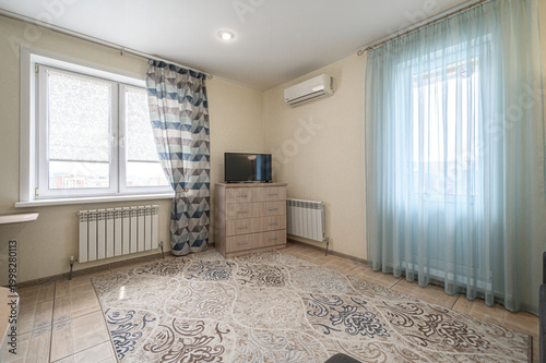 Tablou pe pânză light-filled room with two windows, sheer light blue curtains, patterned rug, wooden dresser with TV, wall-mounted AC, radiators, tiled floor, and geometric-patterned drapes