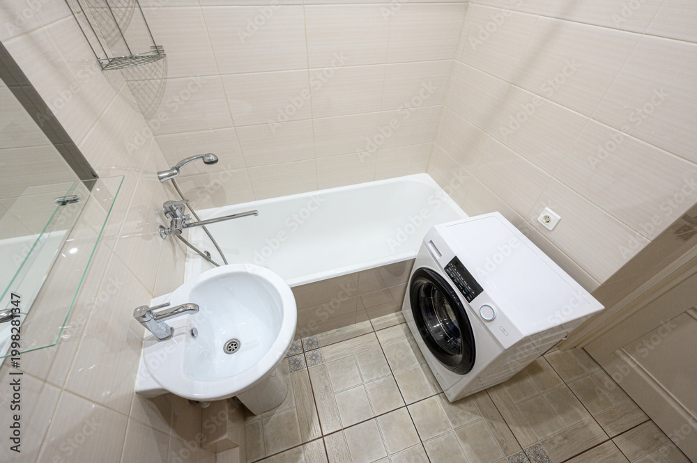 Fototapeta premium compact bathroom with beige tiles, white bathtub, sink on pedestal, toilet, wall-mounted mirror, metal shower rack, washing machine, and grab bar. Clean and functional layout.