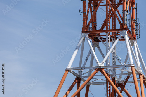 Close-up red-and-white transmission tower under calm blue sky, emphasizing telecommunication industry's infrastructure and showcasing structure's lattice framework, cables, and weathered surface.