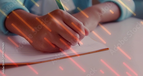 Writing hand on lined orange-bound notebook at desk using green pencil blue cuff orange streaks