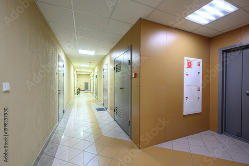 фотографія long, well-lit corridor with beige walls, tiled floor (white yellow), fire safety cabinet, metal fire doors, recessed ceiling lights, and several room doors along the way