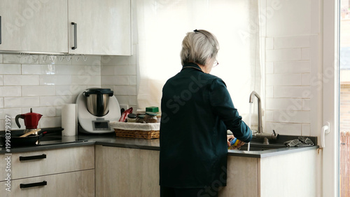 Quadro em tela Senior woman doing daily chores, cleaning kitchen sink with water and sponge