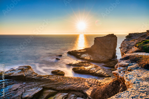 Rocky ocean cliffs with layered stone formations, calm sea, and sunlight reflecting on water at sunset. Scenic coastal landscape with natural textures, warm golden light, and long exposure effect