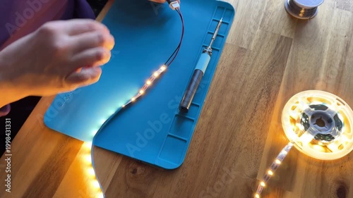 Person solders wires onto LED strip light using soldering iron on blue silicone mat, then tests the glowing warm white LED strip.