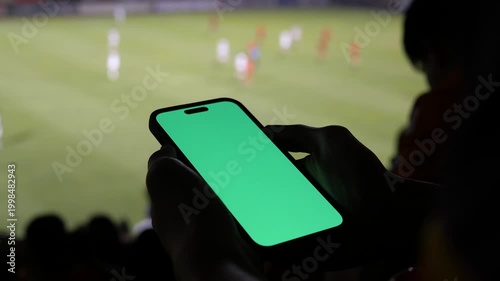 Silhouette hands holding a smartphone with a green screen mockup at a stadium, blurred live soccer match in the background, sports betting and live streaming concept