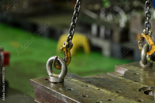 Canvastavla Yellow chain hook locks into steel lifting eye above heavy die block, supporting