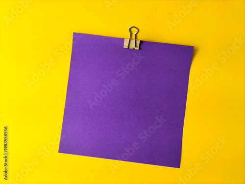 A purple square paper is clipped to a bright yellow background using a metal binder clip. 