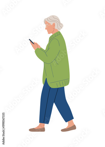 An elderly woman walks while using her phone. She wears a green coat and blue pants. Her hair is short and silver. She walks forward with calm focus. Her expression shows quiet concentration