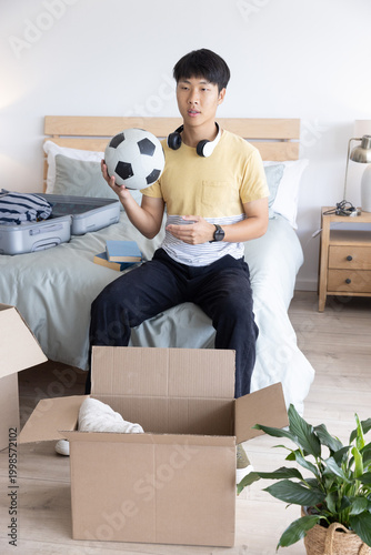 Asian man sitting on edge of bed holding soccer ball while unpacking suitcase boxes and headphones