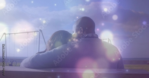 Sitting couple draping arm around woman on wood bench at dusk shore, swinging, bokeh, sweater, coat