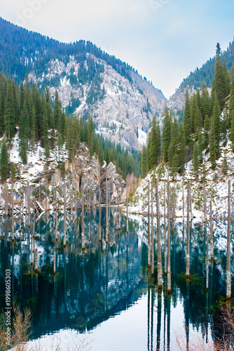 The flooded forest of Lake Kaindy in winter. A beautiful winter mountain landscape in Kazakhstan. A blue lake with tree trunks protruding from it against a backdrop of snow-capped mountains.