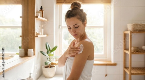 Attractive woman with hair bun applying white body lotion onto her exposed shoulder next to a sunlit window.