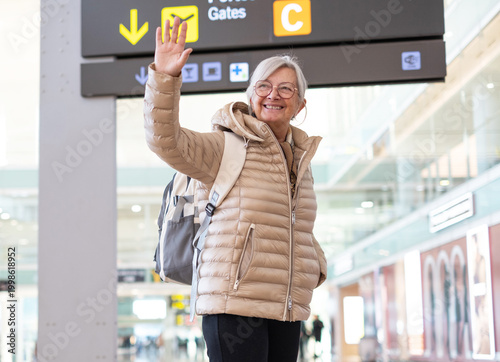 Obraz na płótnie Smiling senior woman waves goodbye in a bright airport terminal wearing a puffer jacket and backpack as she prepares for travel