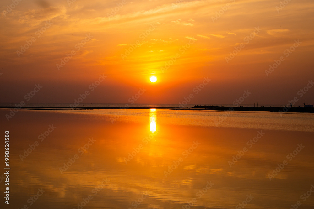 Obraz premium olden sunrise over the sea at Chennai, Tamil Nadu, India. Silhouetted figures stand on a coastal pier as the blazing sun reflects on calm water in a stunning display.