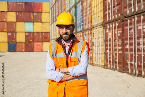 A worker in a yellow hard hat and orange safety vest stands with arms crossed in front of a large stack of colorful shipping containers at a logistics or transportation facility