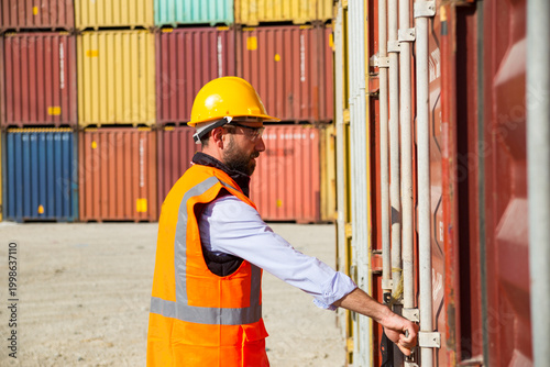 Worker in hard hat and safety vest stands before stacked shipping containers in a port facility