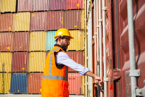 Worker in hard hat and safety vest stands before stacked shipping containers in a port facility