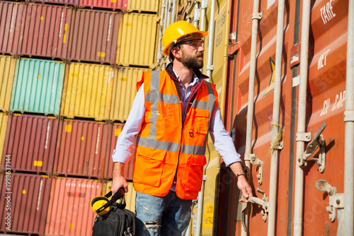 Man in yellow hard hat and orange safety vest standing beside stacked shipping containers at a port