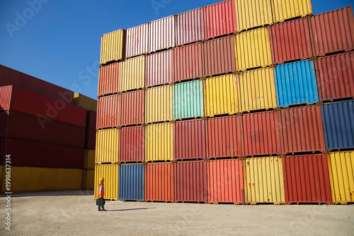 Man in yellow hard hat and orange safety vest standing beside stacked shipping containers at a port