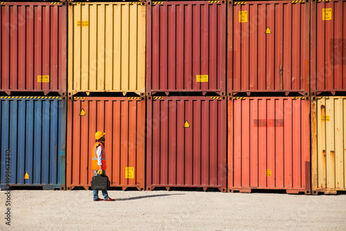 Man in yellow hard hat and orange safety vest standing beside stacked shipping containers at a port