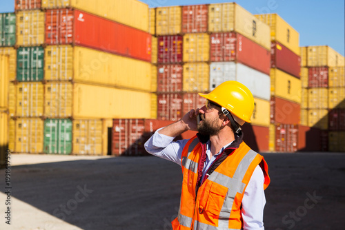 Construction worker in hard hat and safety vest operates radio at shipping container yard