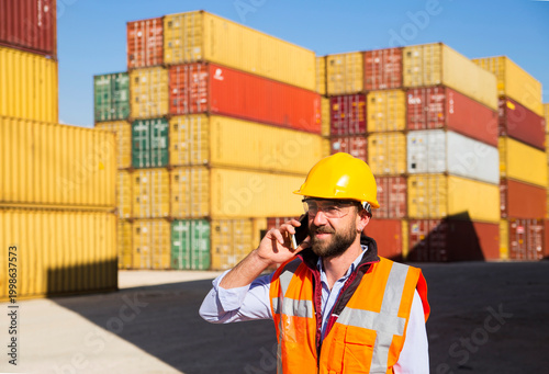 Construction worker in hard hat and safety vest operates radio at shipping container yard