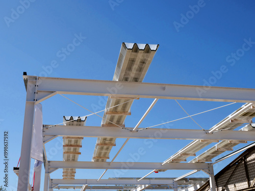  A white steel structural framework for a large building stands
against a clear, bright blue sky. Several long sections of corrugated metal
roofing panels are positioned atop the beams, ready 