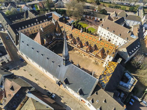 Beaune, France - 05 April 2026: Aerial view of Hotel-Dieu Museum - Hospices de Beaune with its iconic polychrome glazed tile roof and central courtyard under bright sunlight.