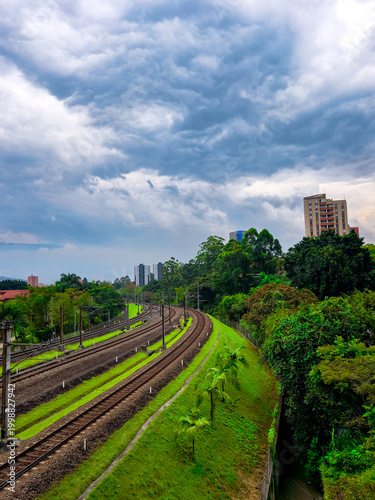 Bild auf Leinwand Curving railway tracks pass through a vibrant green embankment beside dense city vegetation in Medellin, Antioquia, with tall buildings in the distance under a dramatic cloudy sky