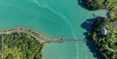 Aerial view of a car crossing a bridge over turquoise water in Kawhia harbour, New Zealand. The road winds along the coast, connecting lush green forests. The image captures travel and nature.