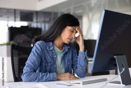 Fényképezés Woman sits at desk, touches her forehead with eyes closed, feels frustrated, overwhelmed or stressed while working at her computer in modern workspace