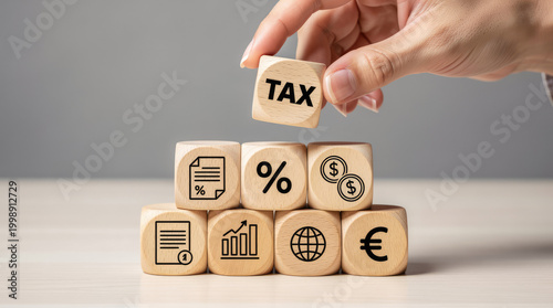 Hand placing a wooden block labeled with the word tax on of a pyramid structure made of cubes with various financial and symbols on a table