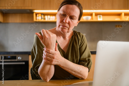 Photos Mature woman massaging sore wrist near laptop at home workspace, discomfort from