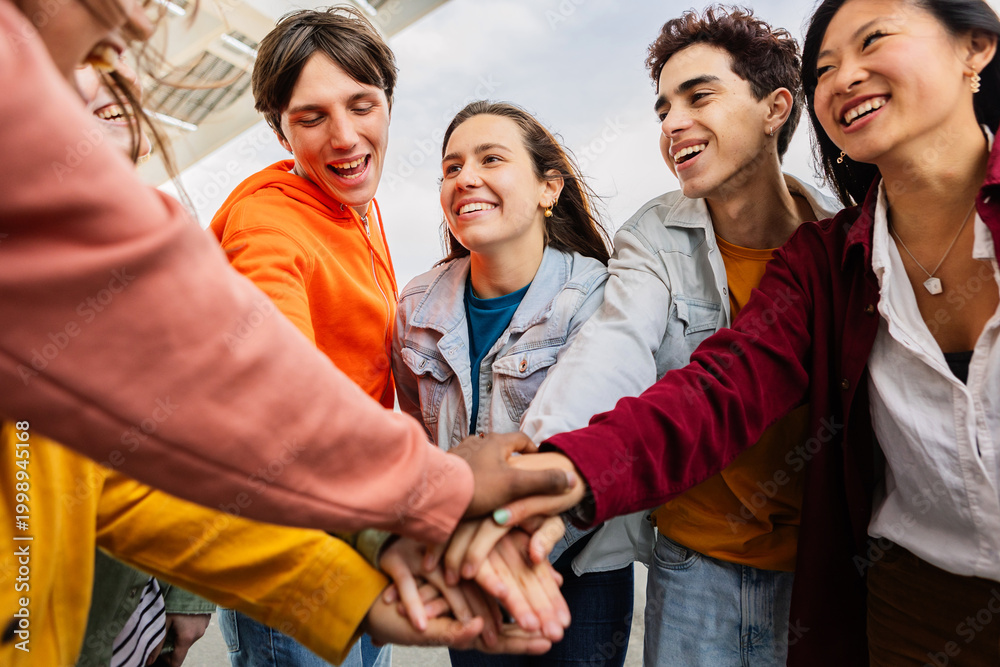 Fototapeta premium Young happy group of diverse people stacking hands outdoors. Multiracial student friends having fun together at city street. Youth community and support concept