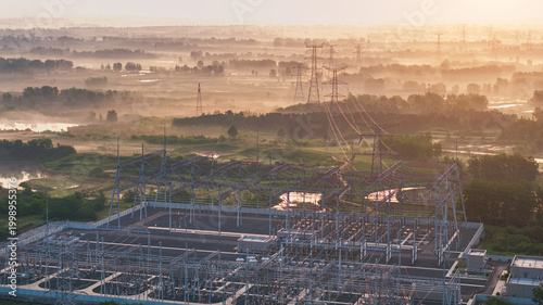 Aerial view of a high voltage substation and transmission towers at misty dawn, with rural wetlands and open fields under warm morning sunlight.