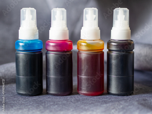 Four refill bottles of printer ink in cyan, magenta, yellow, and black colors arranged in a row on a dark fabric background. High-resolution close-up image suitable for themes related to printing