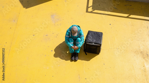 Gig economy worker struggle Top down aerial view of exhausted courier resting against vibrant yellow concrete