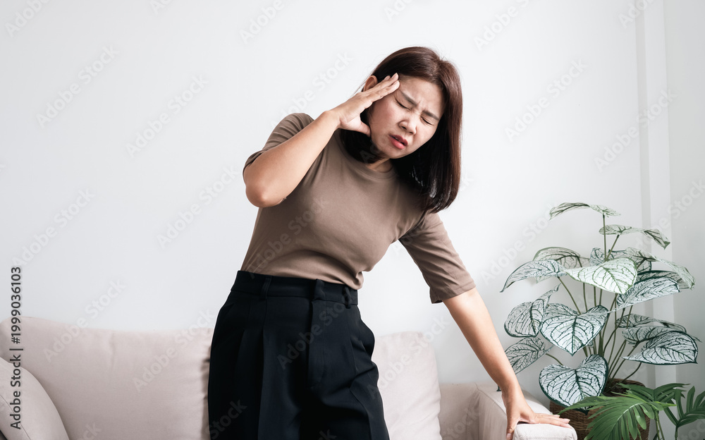 Naklejka premium Asian woman feeling dizzy and faint, holding her head and leaning on sofa for support. Concept of vertigo, hypertension, morning sickness, or low blood pressure. Healthcare and medical theme