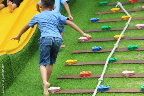 A child is climbing up a wooden obstacle course with colorful grips and a safety rope on green artificial grass. Concept of childhood physical activity, outdoor fitness, and gross motor skills develop