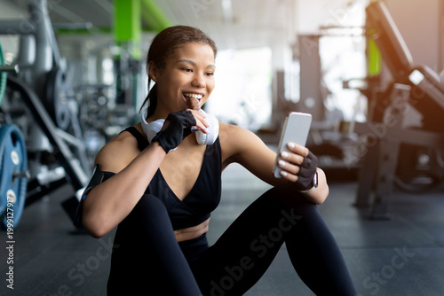 Schilderij op canvas Young Black woman sitting on gym floor eating snack bar and using smartphone, sm