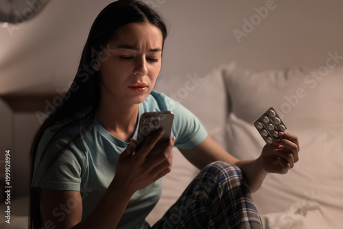 Fotografie Young woman with melatonin pills using mobile phone in bedroom at night