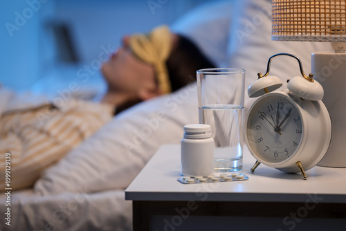 Фотография Melatonin pills with glass of water and alarm clock on table against sleeping wo