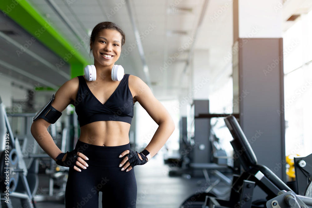 Fototapeta premium Confident smiling young Black woman standing in gym with hands on hips, wearing black sports bra, white headphones, fingerless gloves and phone armband, bright modern gym background