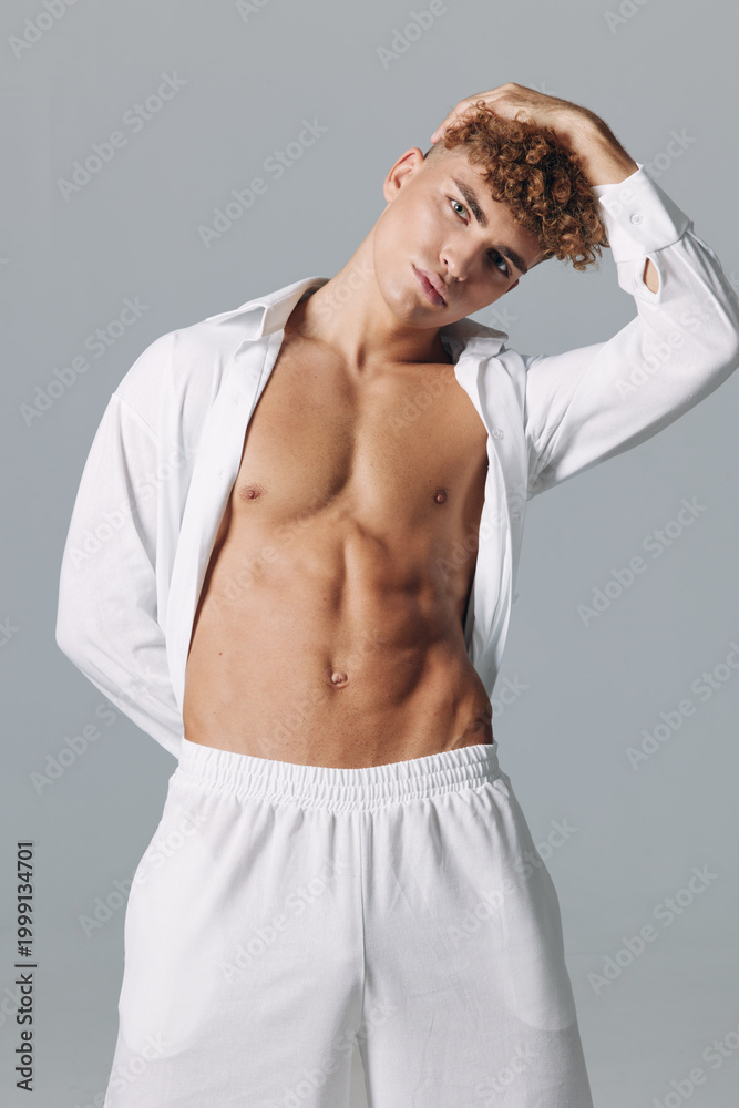 Naklejka premium Young man with curly hair and bare torso posing in white open shirt and shorts cleaning sweat underarm featuring athletic body on neutral studio background close up portrait.