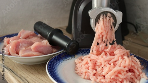 A black electric meat grinder on a kitchen countertop against a gray wall with two plates of pink poultry fillets and ready-made minced meat, minced on a household appliance