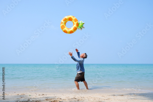 An elderly man joyfully tosses a swim ring by the calm seaside.
