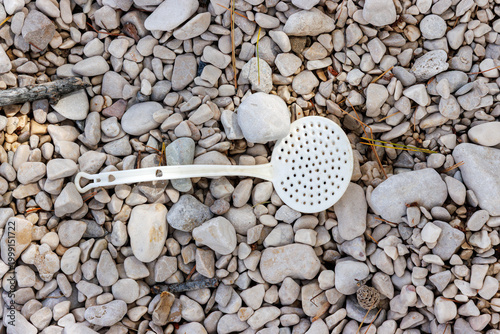Discarded slotted spoon on pebble beach background top view. Plastic waste and beach pollution with copy space.
