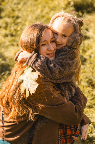 Heartwarming moment: loving mother hugging her little daughter wearing earmuffs in beautiful autumn forest.